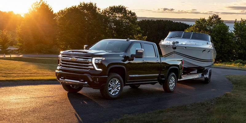 A dark Chevrolet Silverado truck towing a white boat on a trailer, on a paved road at sunset.