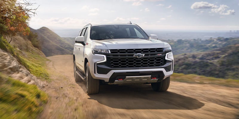Front exterior view of a white Chevrolet Tahoe Z71 driving on a dusty dirt road in the mountains.
