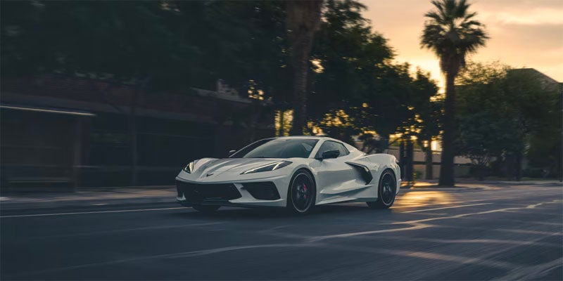 White Corvette C8 Stingray driving on a street at dusk, palm trees and buildings blurred in the background.