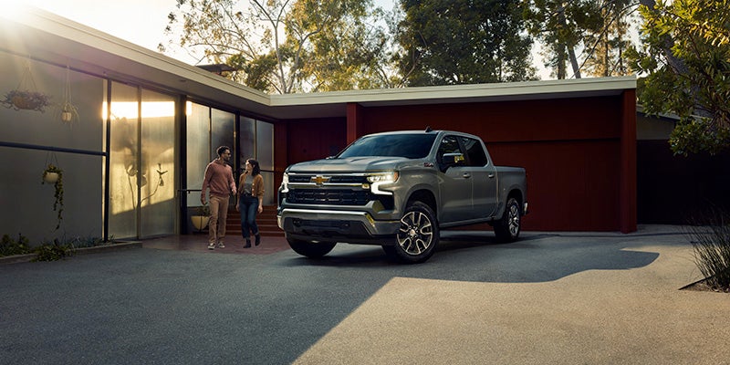 A grey Chevrolet Silverado truck parked outside a modern house with a couple walking in the driveway.