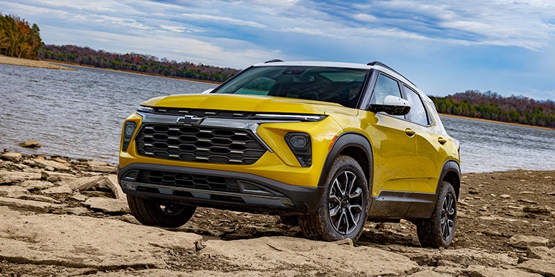 A bright yellow and white Chevrolet Trailblazer SUV parked on a rocky shoreline next to a lake with trees in the distance.